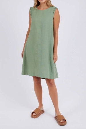 A woman in the Foxwood Bastia Dress, a sleeveless green linen piece with ruffled shoulders, stands against a white background. She pairs it with tan slip-on sandals. Her loose blonde hair perfectly complements the elegant dress.