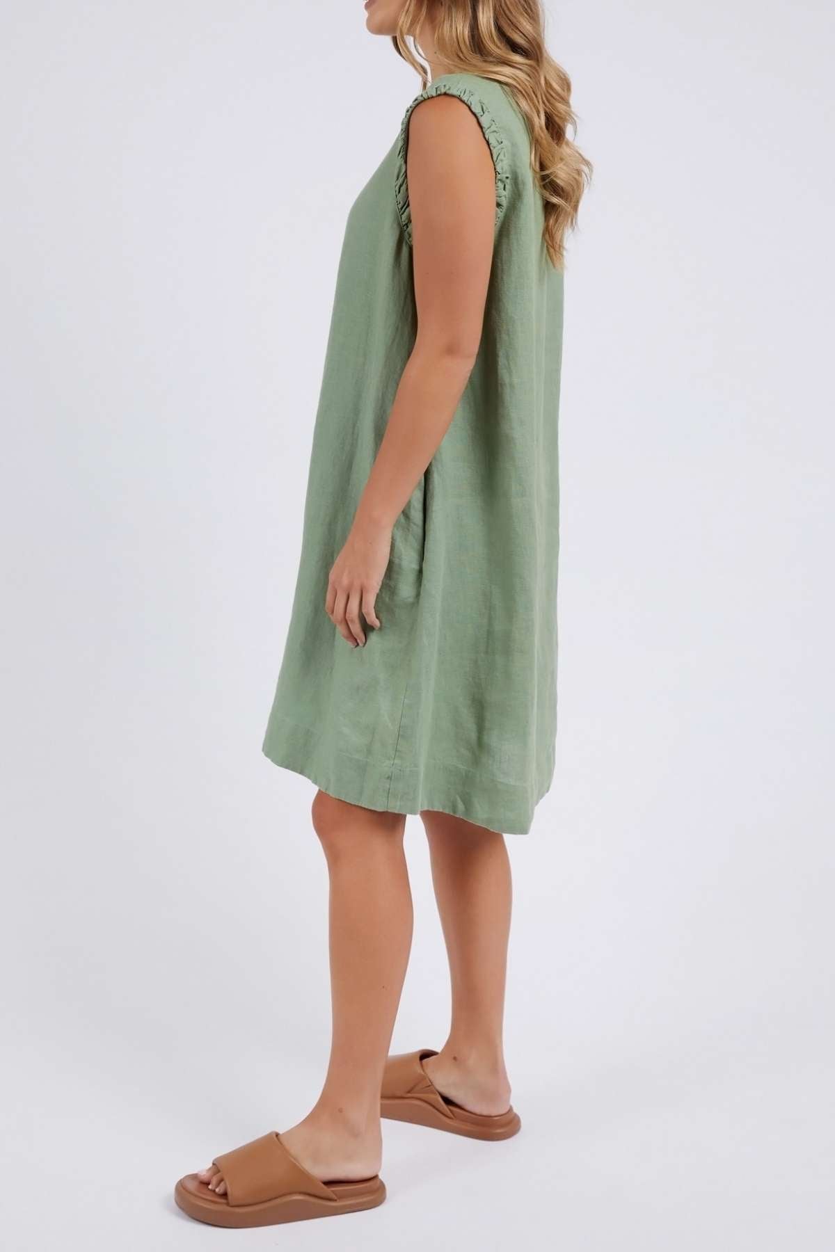 A woman with long wavy hair stands sideways wearing a sleeveless green Foxwood Bastia Dress, paired with tan slide sandals against a plain white background, her hands resting at her sides.