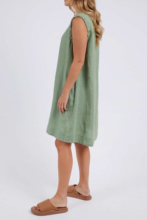 A woman with long wavy hair stands sideways wearing a sleeveless green Foxwood Bastia Dress, paired with tan slide sandals against a plain white background, her hands resting at her sides.