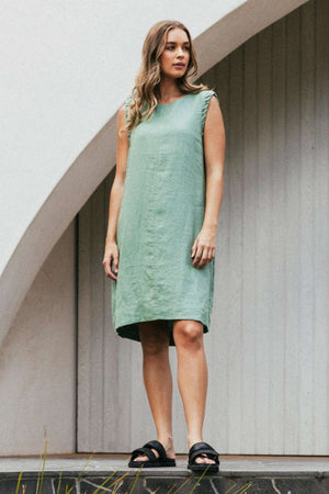 Dressed in the Foxwood Bastia Dress, a woman with long, wavy hair and black slide sandals stands by an arched doorway, exuding chic elegance in the sleeveless green linen.