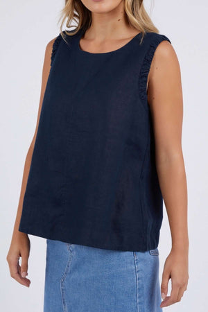 A woman wears the Foxwood Bastia Top—a relaxed-fit, sleeveless navy top with ruffled shoulders—paired with a light blue denim skirt. The photo is cropped at her chin, focusing on her upper body.