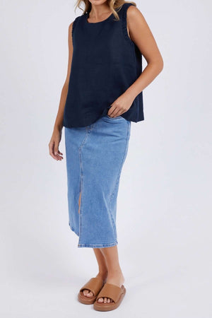 Wearing a relaxed fit Foxwood Bastia Top in navy, a knee-length denim skirt with a front slit, and tan slip-on sandals, the person stands against a plain white background.