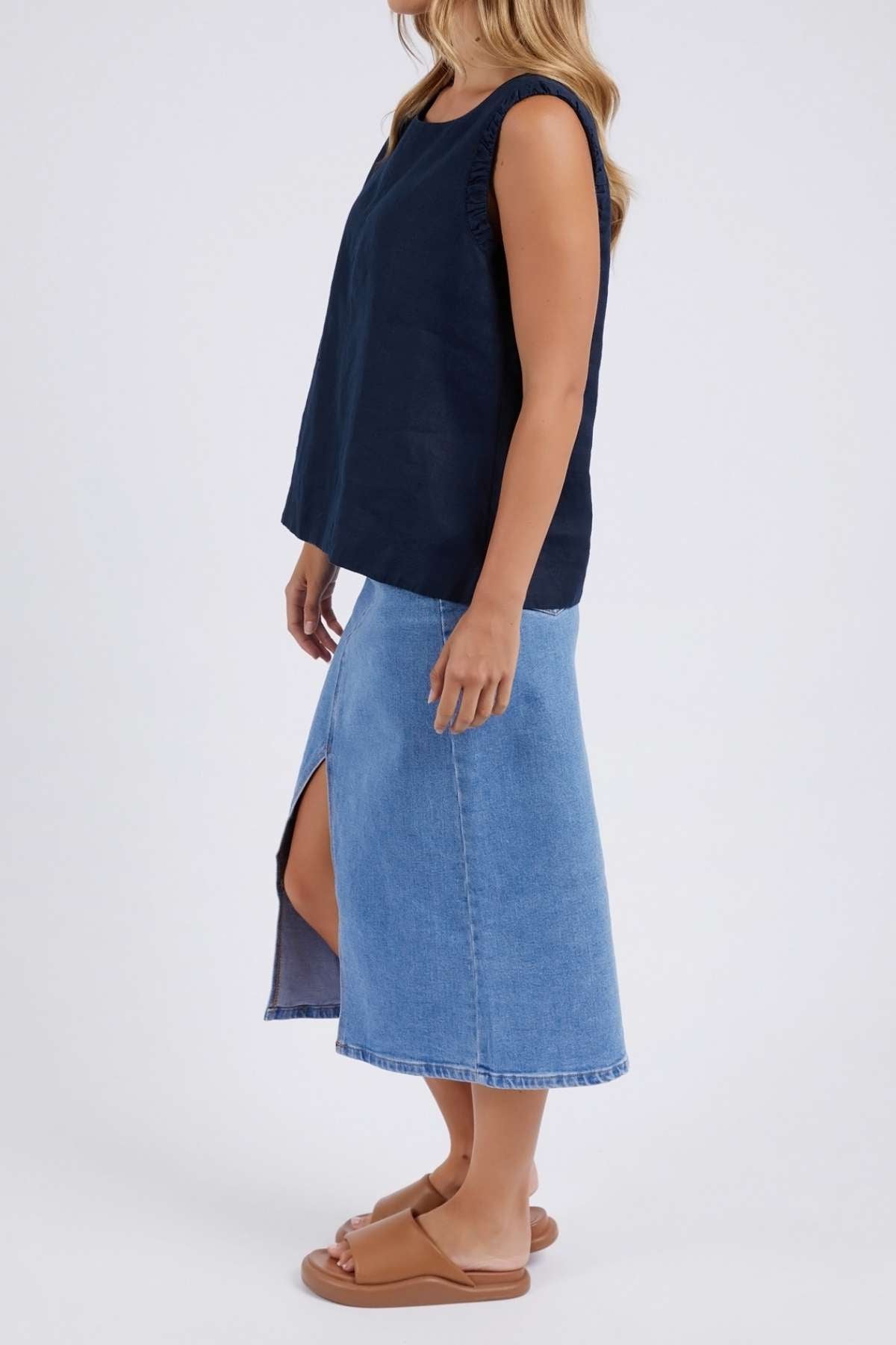A woman wears the Foxwood Bastia Top in navy, a relaxed-fit 100% linen piece, paired with a light blue denim midi skirt with front slit and tan sandals, standing against a plain light background.