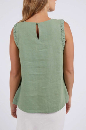 A stylish woman stands facing away, wearing a green sleeveless Foxwood Bastia Top with ruffled shoulders and a keyhole button closure, paired with a white skirt against a plain light backdrop.