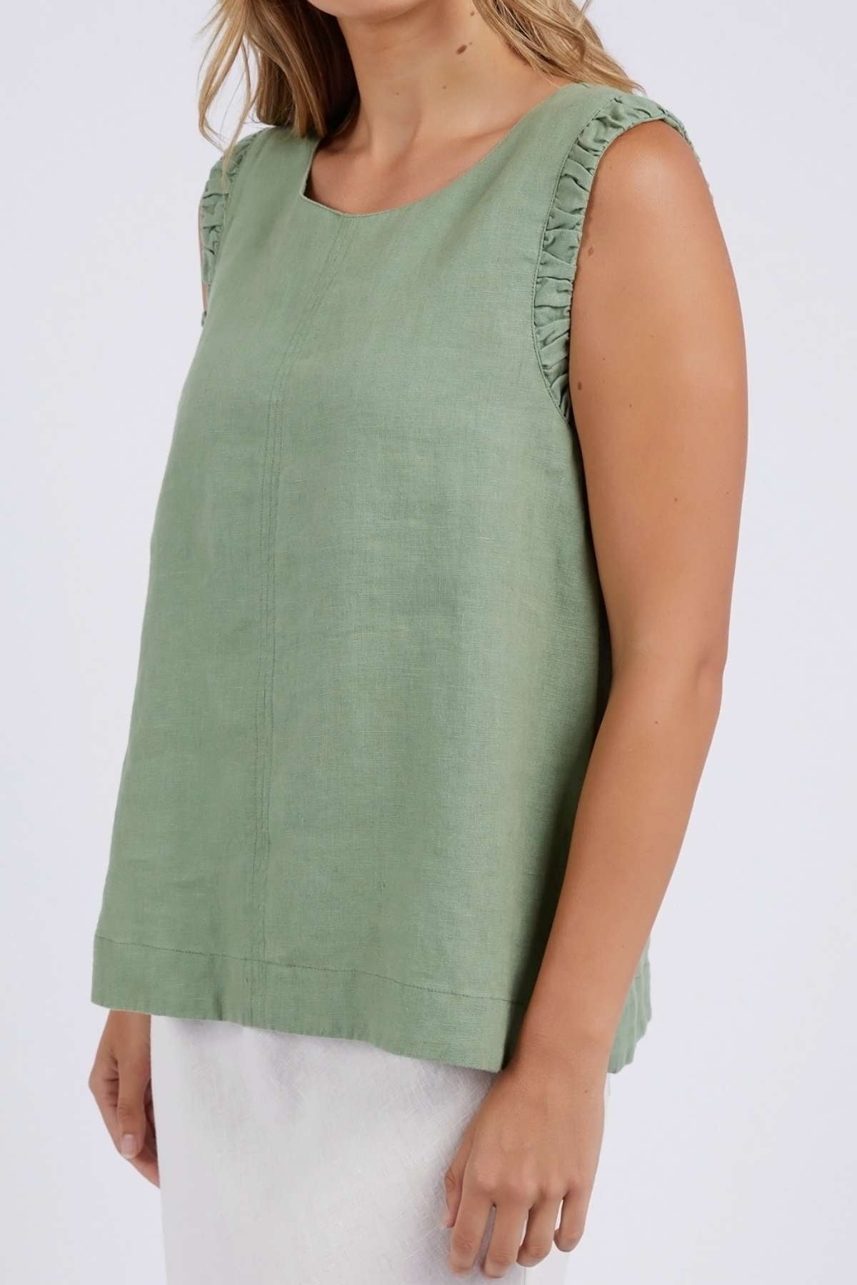 Wearing a sleeveless, loose-fitting green Foxwood Bastia Top with ruffled shoulders, they present a fashion-forward vibe. Their light wavy hair and the white skirt complement the ensemble against a plain background. The image is cropped at the neck and knees.