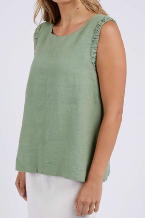 Wearing a sleeveless, loose-fitting green Foxwood Bastia Top with ruffled shoulders, they present a fashion-forward vibe. Their light wavy hair and the white skirt complement the ensemble against a plain background. The image is cropped at the neck and knees.