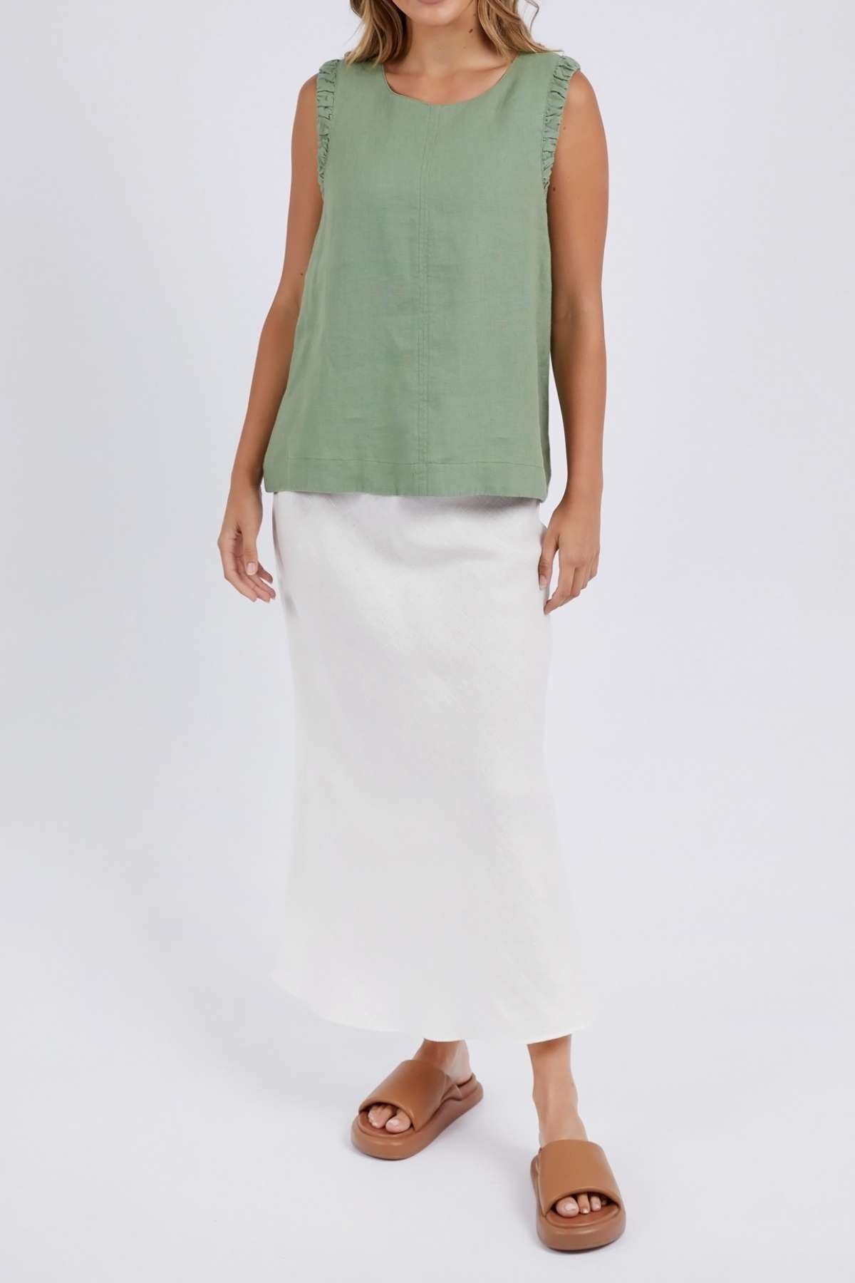 Dressed in a trendy Foxwood Bastia Top and a long white skirt, the individual poses against a simple backdrop, wearing brown slide sandals. Their face is hidden from view.