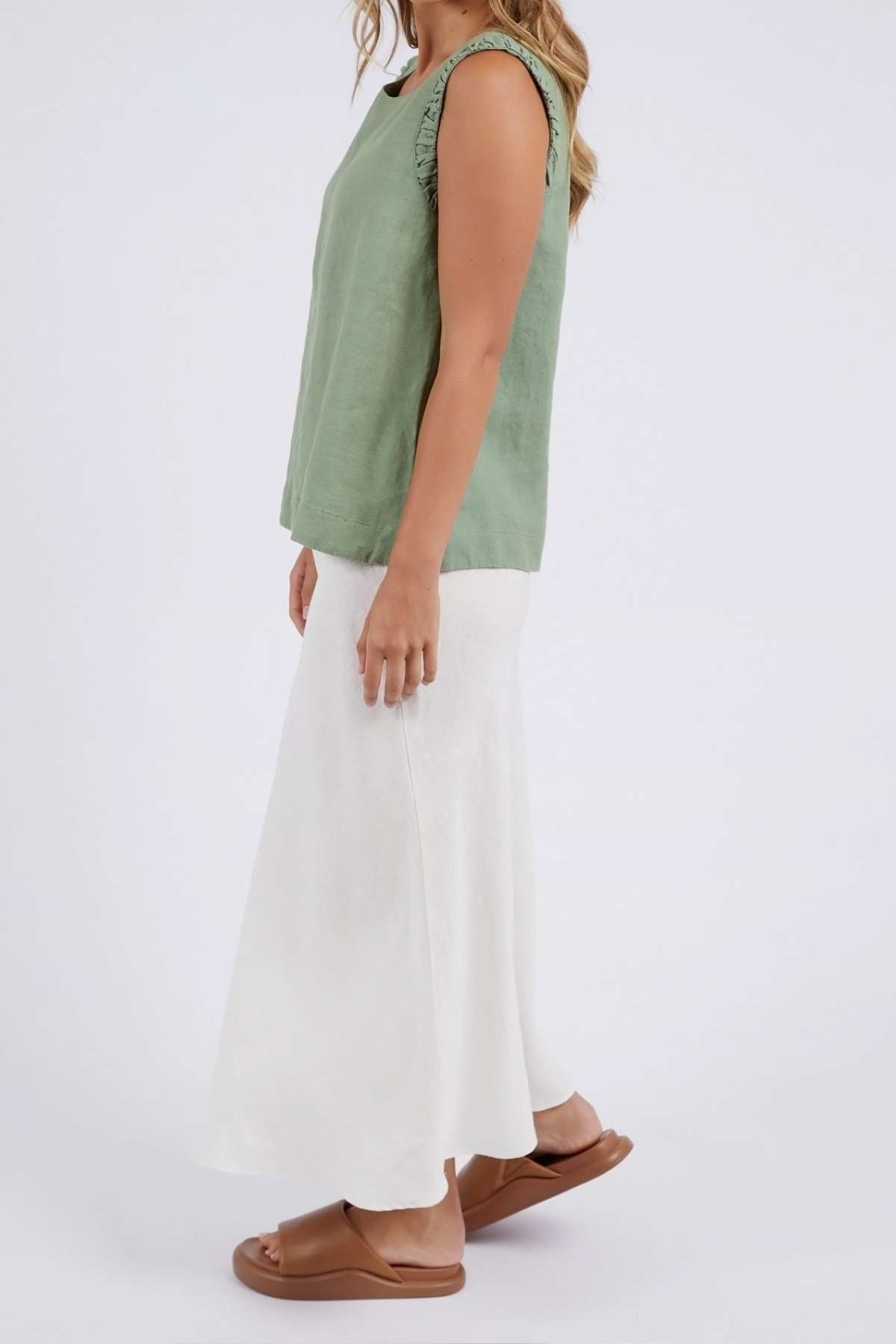 A woman stands sideways in a sleeveless, fashion-forward Foxwood Bastia Top and a flowing white skirt. She pairs her outfit with brown slide sandals, long hair cascading down her back, creating an effortlessly chic look against the plain white background.
