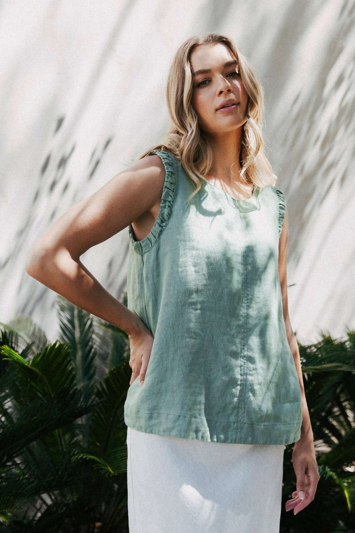 A person with long blond hair is outdoors, donning a sleeveless Foxwood Bastia Top and a white skirt. Greenery envelops them as shadows dance on the luminous backdrop, creating an effortlessly chic ensemble.
