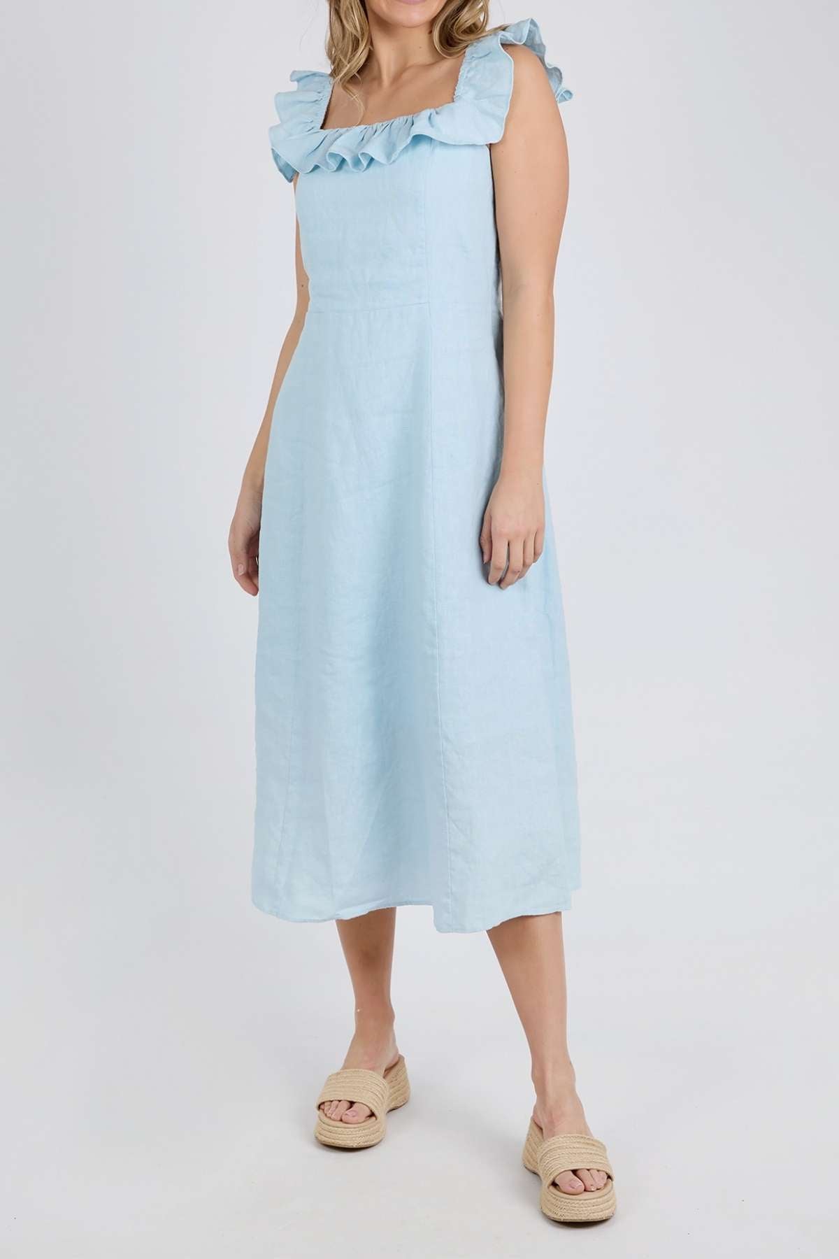A woman models the Foxwood Bonifacio Dress in light blue linen with ruffled straps, paired with beige platform sandals, standing against a plain white background. Her face is not shown.