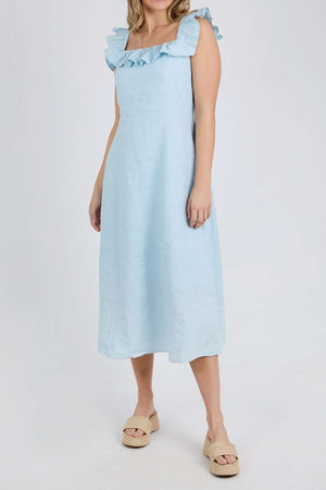 A woman models the Foxwood Bonifacio Dress in light blue linen with ruffled straps, paired with beige platform sandals, standing against a plain white background. Her face is not shown.