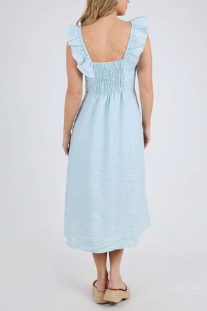 A person models the light blue Foxwood Bonifacio Dress, featuring luxe linen, ruffled straps, and a smocked back, styled with beige platform sandals.