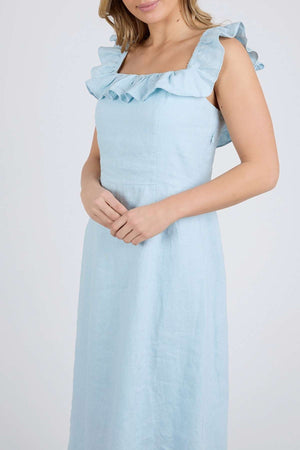 A woman models the Foxwood Bonifacio Dress, featuring ruffled straps and crafted from luxe light blue linen, standing against a plain background with her hands clasped at her waist.