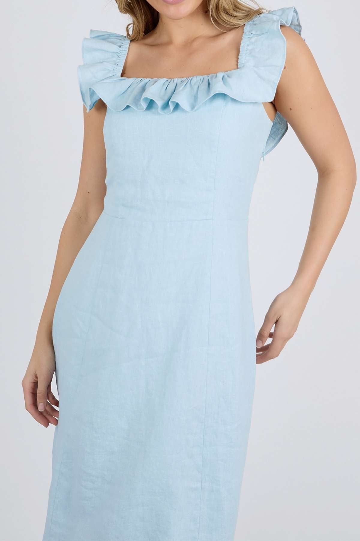 A woman in the Foxwood Bonifacio Dress—a light blue, sleeveless linen-blend style with ruffled square neckline—stands against a plain light background.