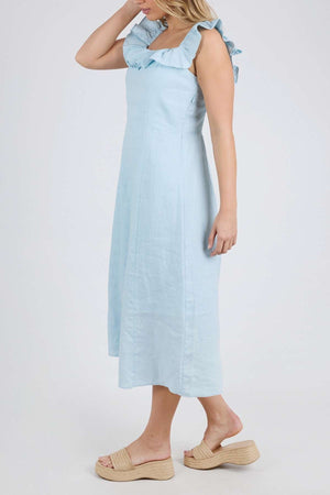 A woman in the Foxwood Bonifacio Dress—a light blue, sleeveless linen midi with ruffle straps—pairs it with beige platform sandals and stands before a plain, light background.