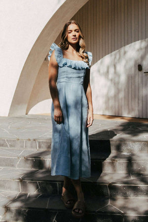 A woman wears the Foxwood Bonifacio Dress in light blue linen, standing on stone steps by a curved white building as sunlight and shadows form patterns on the wall and ground.