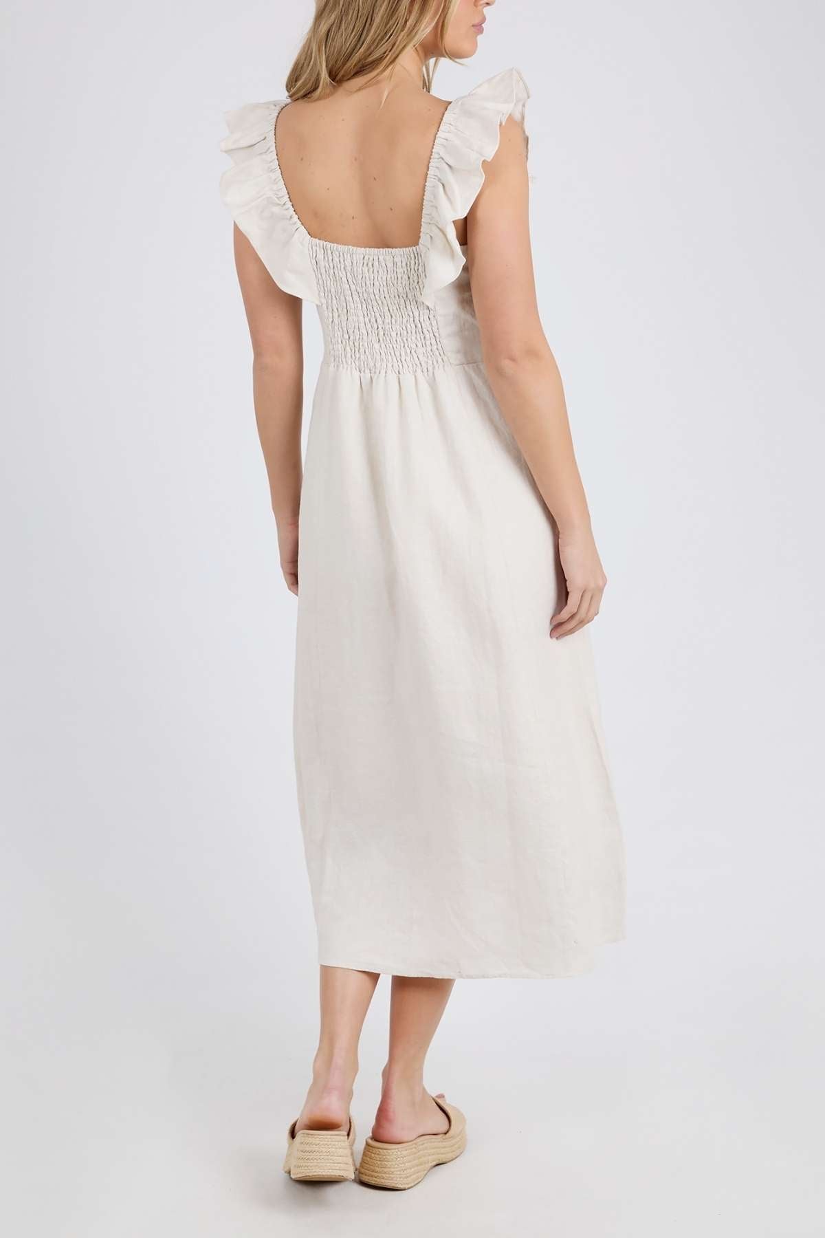 A rear view shows a person with long hair in a white Foxwood Bonifacio Dress, featuring ruffled straps and a shirred back in luxe linen. The dress falls to mid-calf, paired with beige platform sandals against a plain white background.