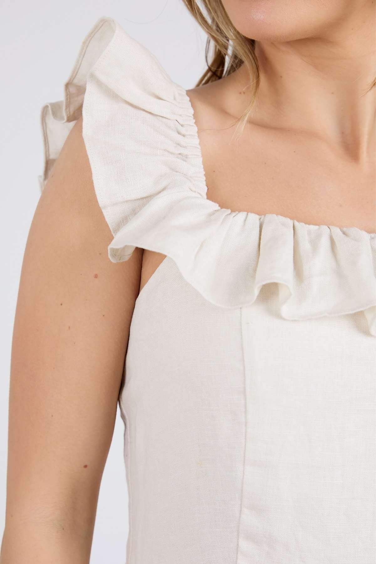 An elegantly dressed woman wears a sleeveless Foxwood Bonifacio Dress, made of luxe linen with ruffled straps. The off-white attire is shown from shoulders to upper torso against a simple backdrop.