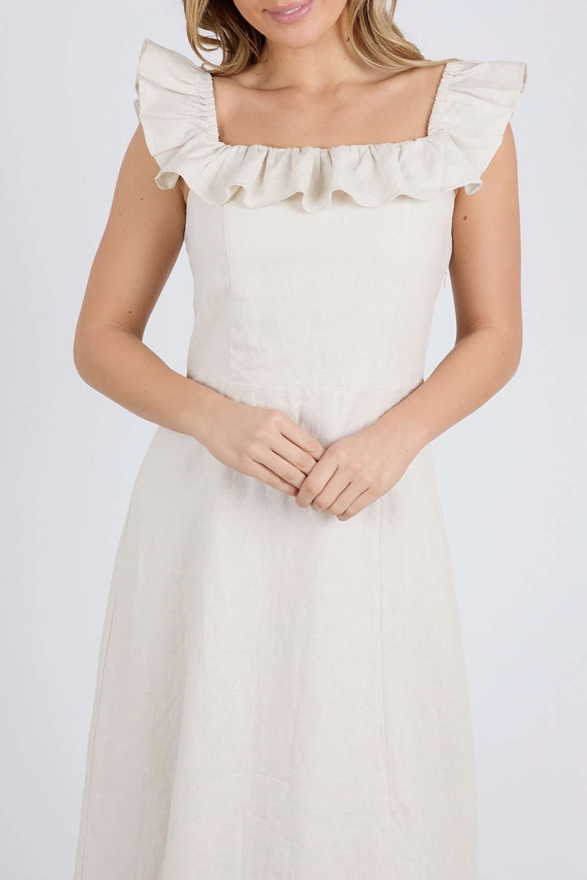 A woman in a cream Foxwood Bonifacio Dress, featuring ruffled shoulders and a square neckline, stands against a plain background holding her hands gently in front of her. The sleeveless, luxe linen dress is simple and elegant.