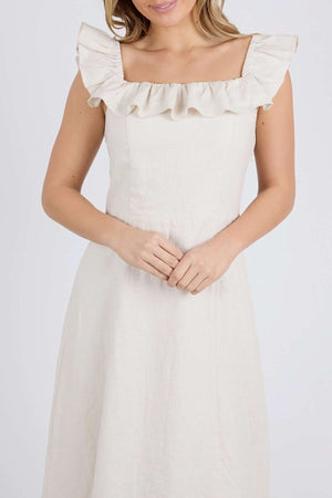A woman in a cream Foxwood Bonifacio Dress, featuring ruffled shoulders and a square neckline, stands against a plain background holding her hands gently in front of her. The sleeveless, luxe linen dress is simple and elegant.