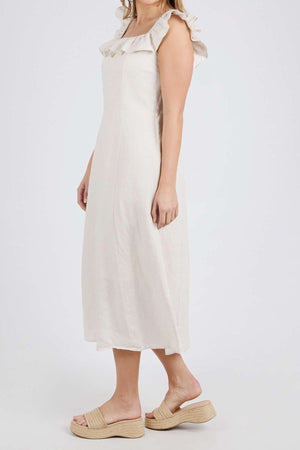 Wearing the luxe linen Foxwood Bonifacio Dress, a sleeveless, ruffled, off-white mid-length piece made from viscose, a person stands sideways paired with woven platform sandals against a plain white background.