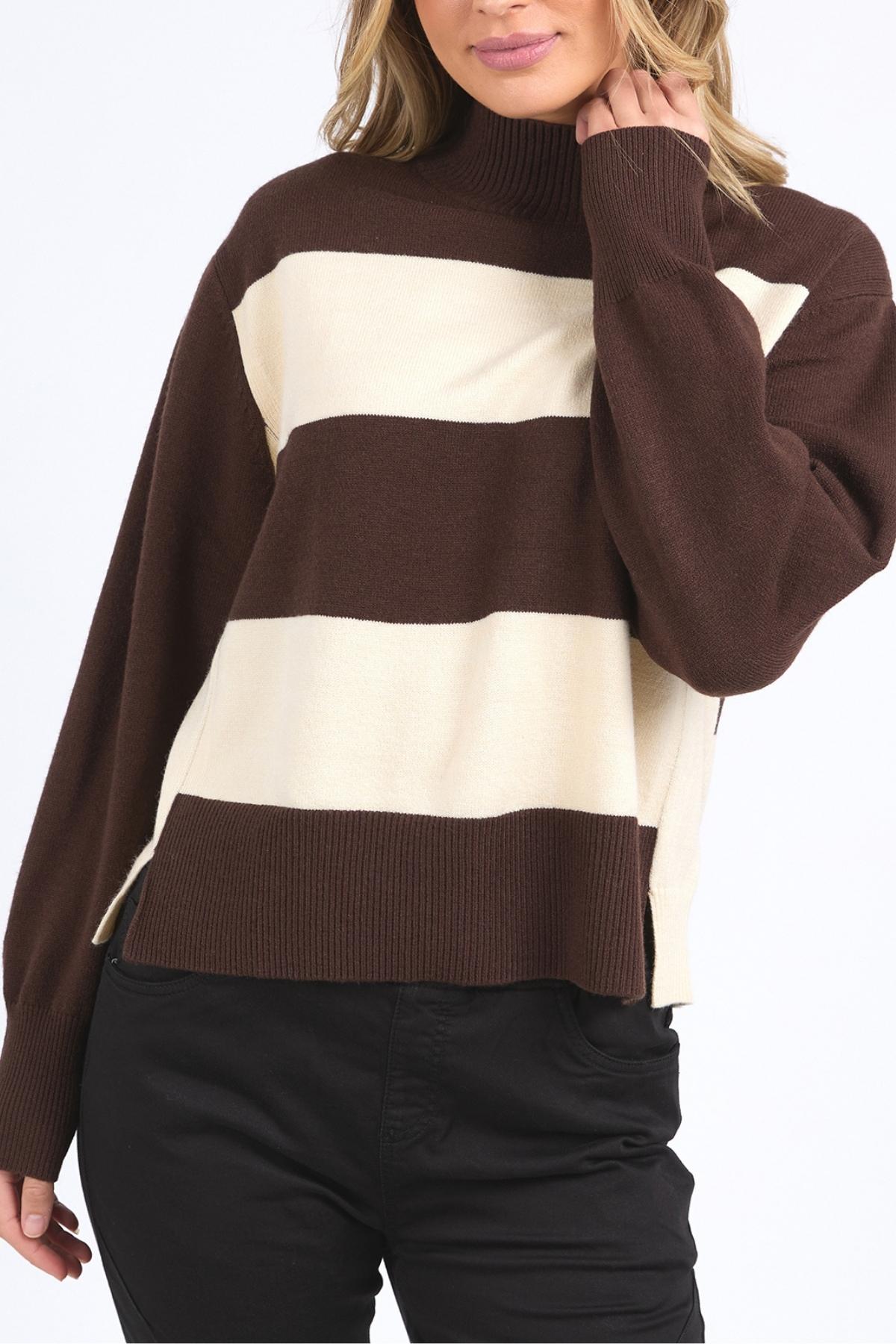 A person wearing the Foxwood Canterbury Knit—a brown and cream yarn-dyed stripe sweater with long sleeves, ribbed cuffs, and a high neck—paired with black pants. Their hair is loose and their hand touches their face.