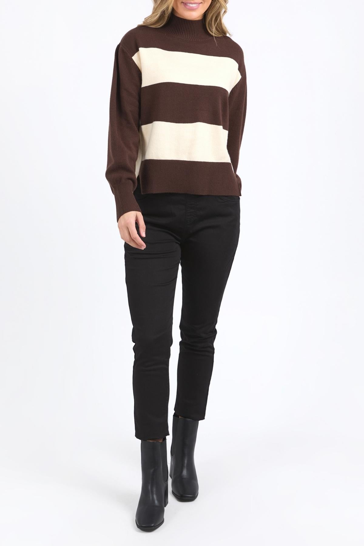 A person wearing the Foxwood Canterbury Knit, featuring brown and cream yarn-dyed stripes, with black pants and black ankle boots stands against a plain white background. The person’s face is not visible.