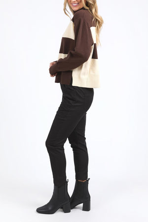 Woman standing sideways, smiling, wearing the Foxwood Canterbury Knit in brown and cream stripes, paired with black skinny pants and black ankle boots against a plain white background.