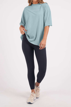 Wearing a Foxwood Captain Tee, an oversized light blue cotton t-shirt, paired with dark leggings and white sneakers. Ideal for casual occasions, they stand against a plain backdrop with one hand in their pocket and one knee slightly bent.