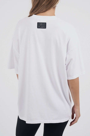 A person with long hair wears the Foxwood Captain Tee—an oversized, loose white cotton T-shirt with a small black and white rectangular logo near the neckline—paired with black pants, shown from the back. Ideal for any casual occasion.