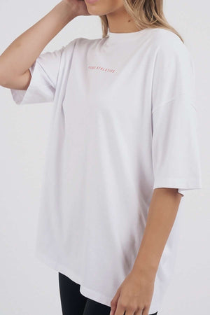 A woman wears the Foxwood Captain Tee—an oversized, loose white cotton t-shirt with short sleeves and subtle light pink chest text. With light brown hair and a plain background, it's perfect for casual occasions.