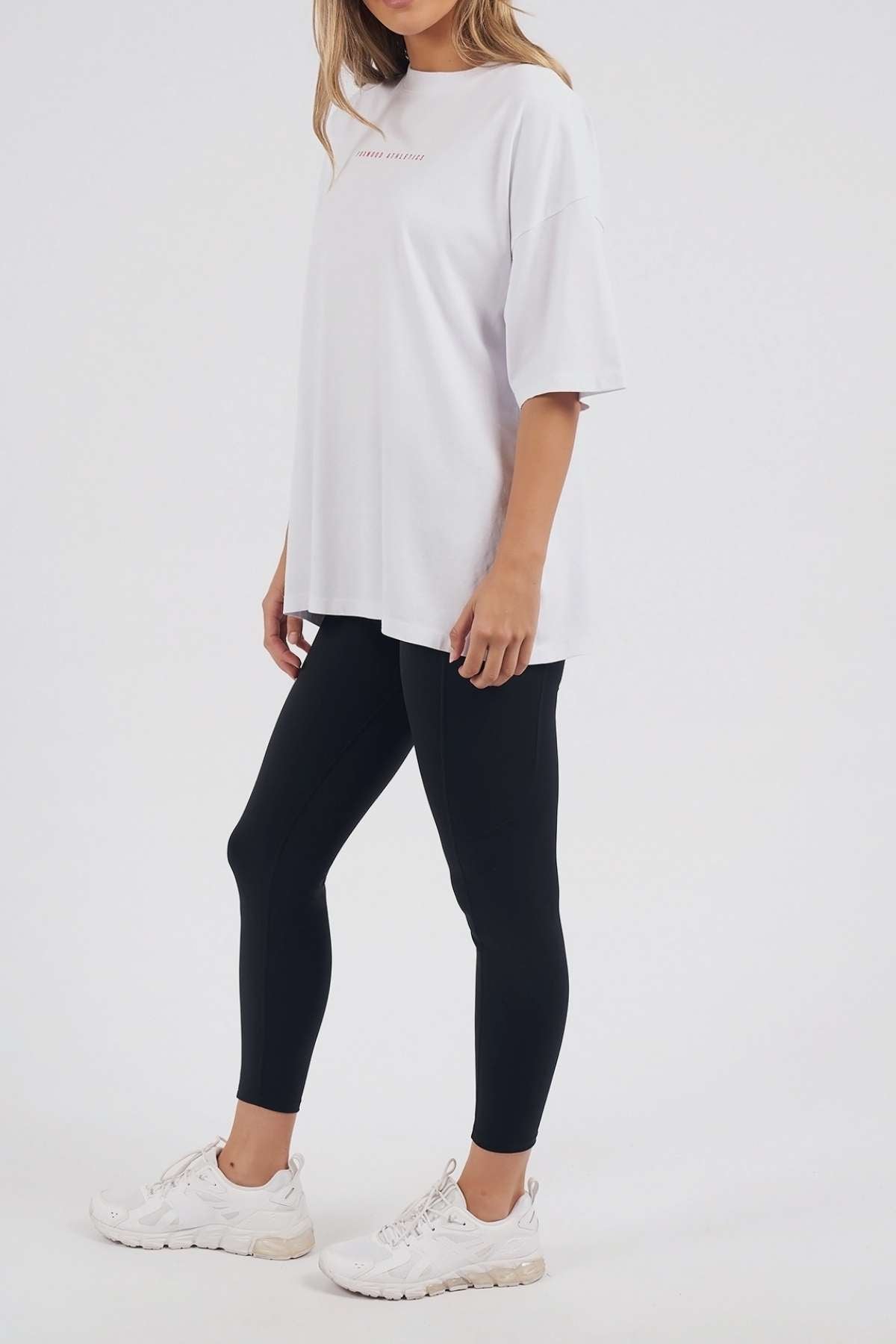 Wearing the Foxwood Captain Tee in white, styled with black leggings and white sneakers, this person stands against a plain light background. Their face is not shown—ideal for a relaxed, casual look.