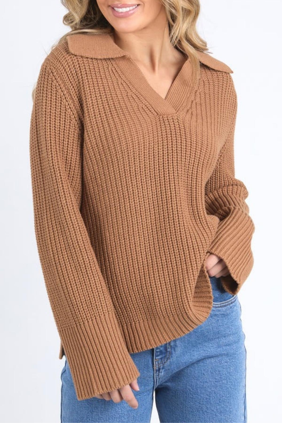 A woman models the Foxwood Carlton Knit, a collared light brown ribbed sweater with wide sleeves, paired with blue jeans. She smiles and stands against a plain white background, highlighting the sweater’s comfortable and stylish look.