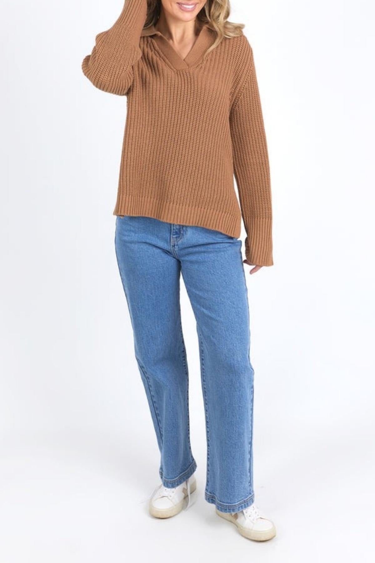 Wearing the Foxwood Carlton Knit with blue jeans and white sneakers, a person stands against a white background. Their relaxed-fit knit exudes comfort and style. One hand is raised near their head, and their face remains unseen.