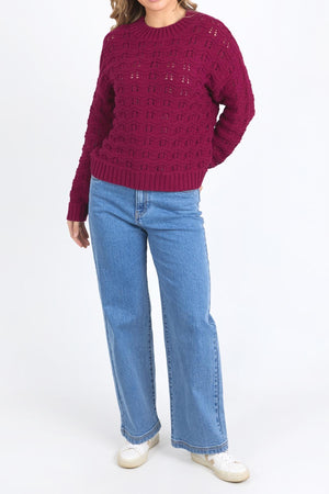 A person in a maroon Foxwood Carter Knit sweater, blue wide-leg jeans, and white sneakers stands against a plain white background, with their head partially out of frame.