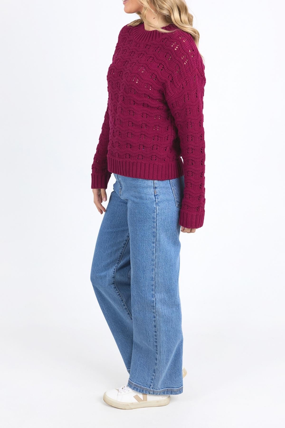 A person wears the maroon Foxwood Carter Knit sweater, made from a cotton-acrylic blend, paired with light blue wide-leg jeans and white sneakers, standing against a plain white background slightly turned to the side.