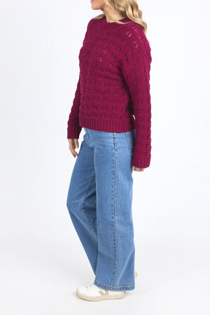 A person wears the maroon Foxwood Carter Knit sweater, made from a cotton-acrylic blend, paired with light blue wide-leg jeans and white sneakers, standing against a plain white background slightly turned to the side.