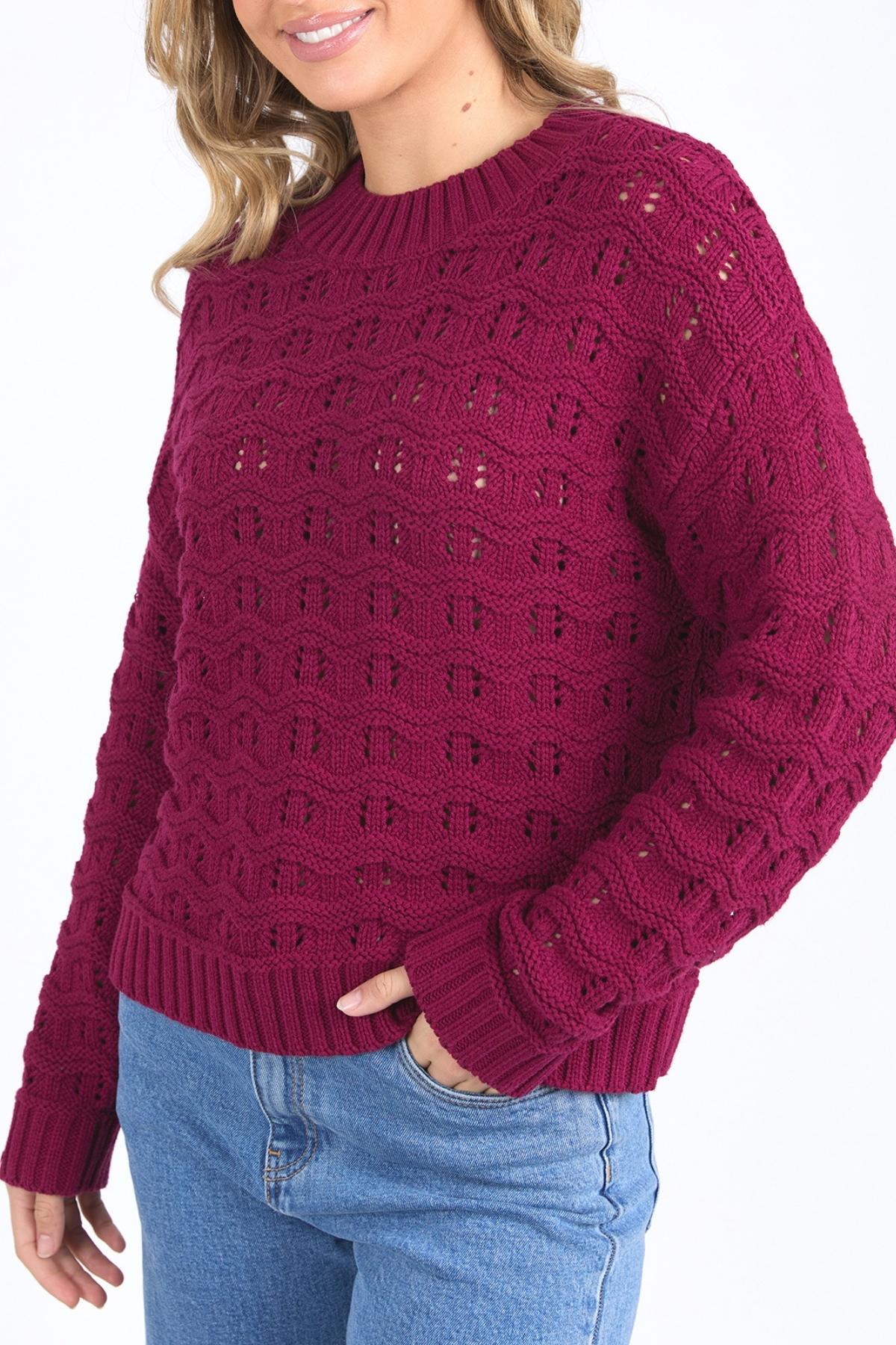 A woman smiles slightly, standing with one hand in her pocket, wearing blue jeans and a textured Foxwood Carter Knit sweater in dark red. The sweater features ribbed hem and cuffs with a subtle pattern.