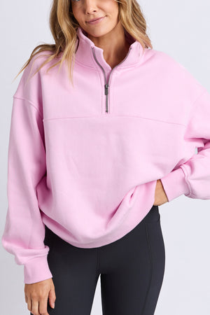 A woman stands against a plain background wearing the Foxwood College Contrast Zip Crew in light pink, a relaxed-fit 100% cotton jumper, paired with black leggings, with one hand in her pocket.