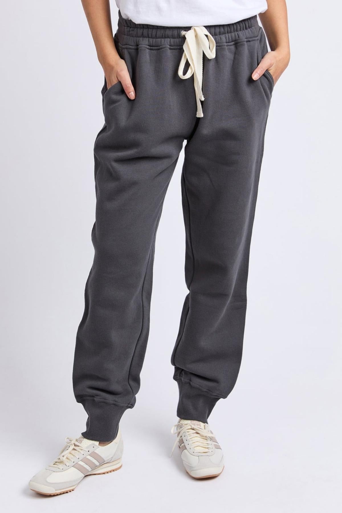 Person wears Foxwood College Track Pant, a dark gray jogger with elastic waist and off-white drawstring, paired with a white tee and white sneakers featuring beige details, standing against a plain light background.