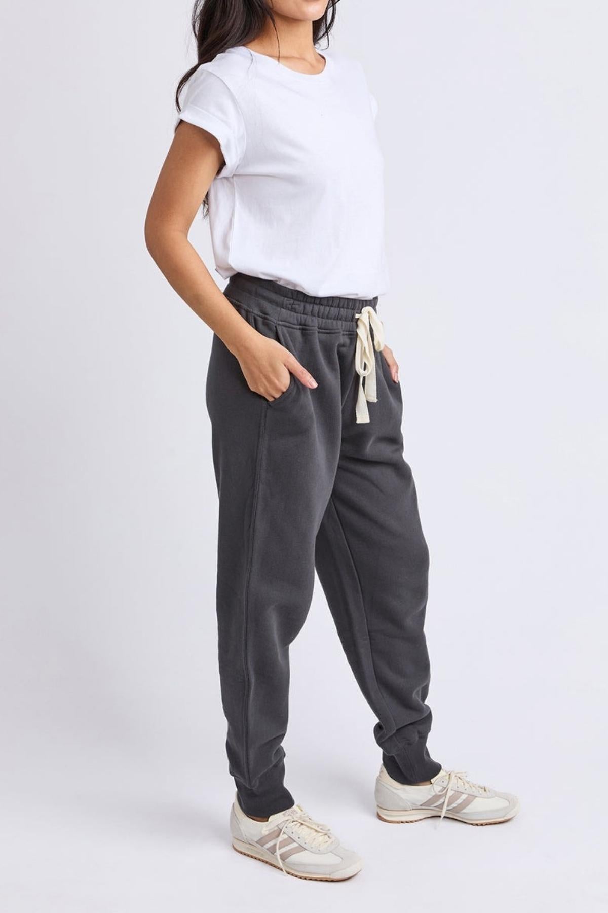 A person stands with hands in pockets, wearing a white t-shirt, Foxwood College Track Pant joggers with an elastic waist and drawstring, and white sneakers against a plain light background.