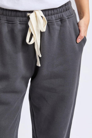Person wears Foxwood College Track Pant in dark gray with elastic waist and white drawstring, hands in pockets, standing against a plain light background. Only lower torso and upper legs are visible, highlighting the timeless silhouette.