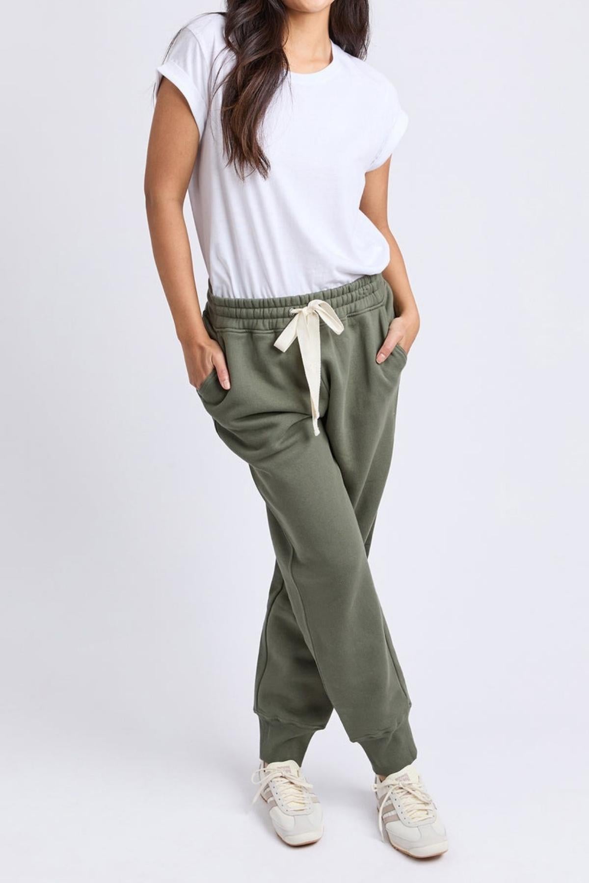 A woman stands with her hands in the pockets of Foxwood College Track Pant, pairing it with a white t-shirt and sneakers, showcasing comfort and style against a plain light background.