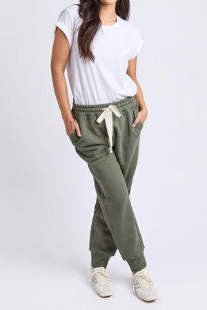 A woman stands with her hands in the pockets of Foxwood College Track Pant, pairing it with a white t-shirt and sneakers, showcasing comfort and style against a plain light background.