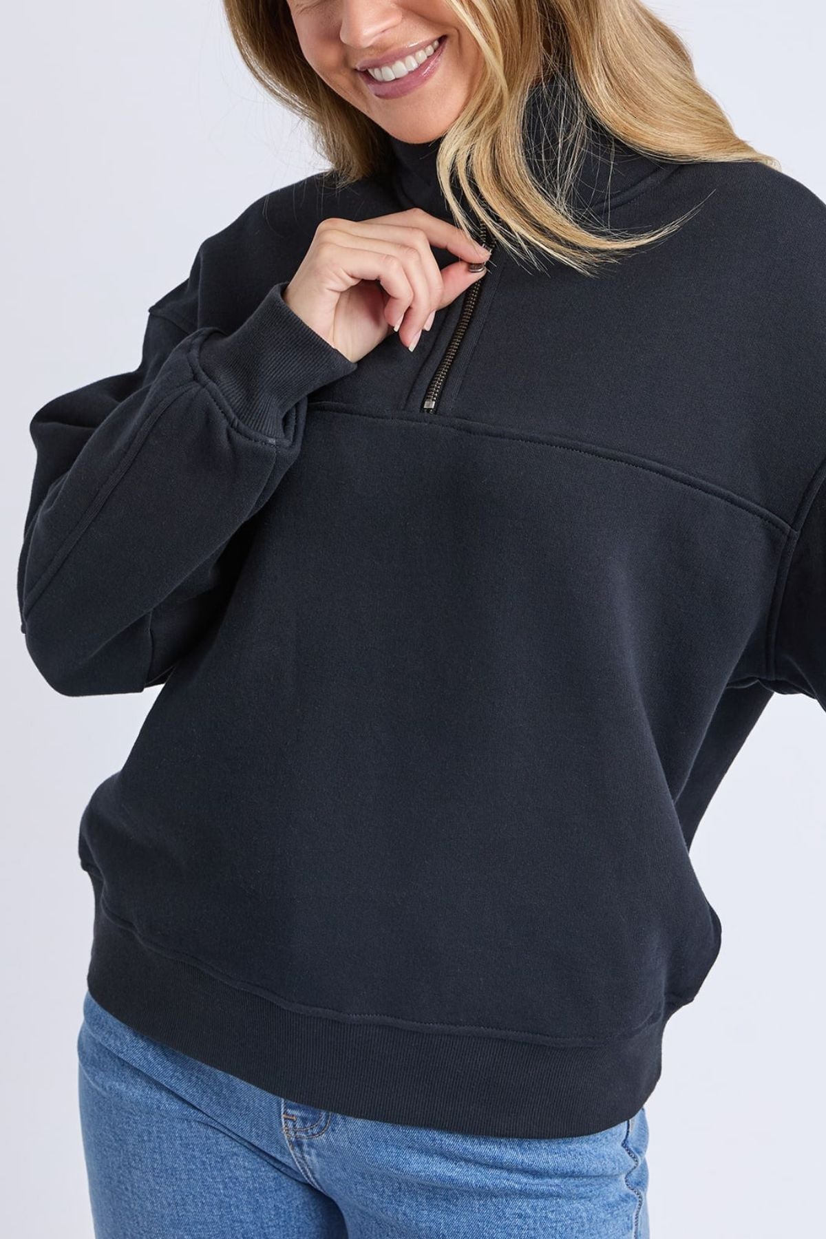 A woman in a relaxed fit Foxwood College Zip Crew sweatshirt (black), paired with blue jeans, smiles as she touches the collar zipper. Her face is partially out of frame.