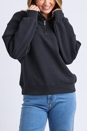 Someone smiles while wearing the Foxwood College Zip Crew, a relaxed fit black sweater with a half-zip and soft cotton feel, paired with light blue jeans and standing against a plain light background.