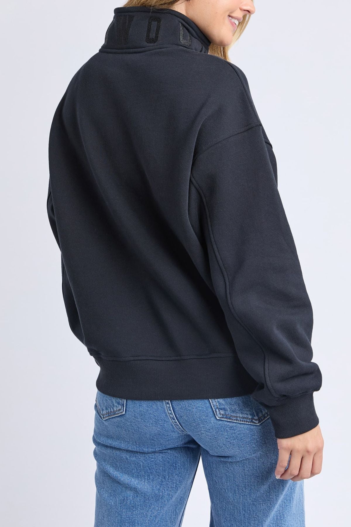 A woman in a black Foxwood College Zip Crew sweatshirt and blue jeans is shown from the back, turning slightly right and smiling against a plain light gray background.