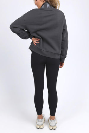 A person faces away in a dark gray Foxwood College Zip Crew with an embroidered collar, black leggings, and white sneakers. One hand rests on their hip against a plain white background. The zip crew features a relaxed fit.