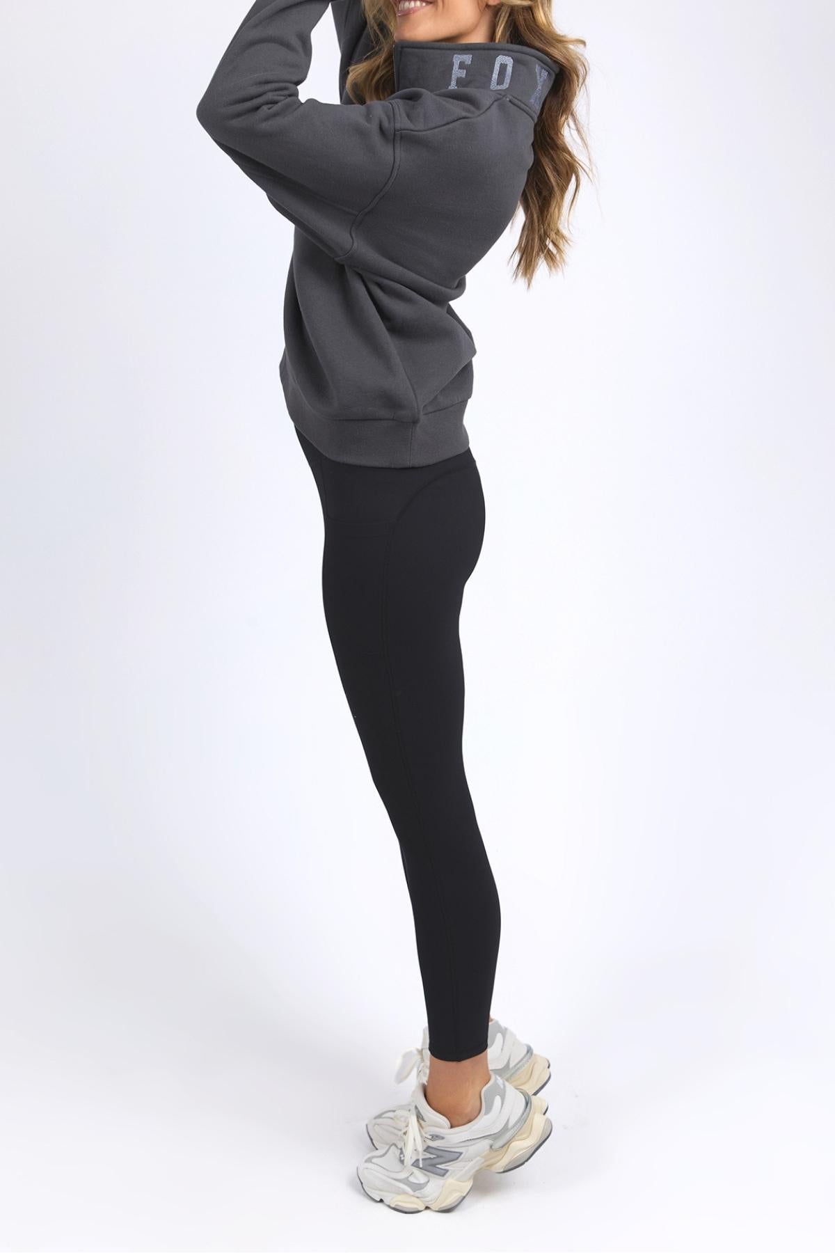 A woman wearing the Foxwood College Zip Crew in dark gray, paired with black leggings and white sneakers, stands sideways on a white background with her long hair loose and hands raised near her face.