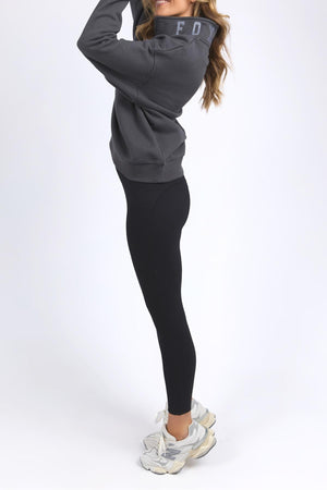 A woman wearing the Foxwood College Zip Crew in dark gray, paired with black leggings and white sneakers, stands sideways on a white background with her long hair loose and hands raised near her face.